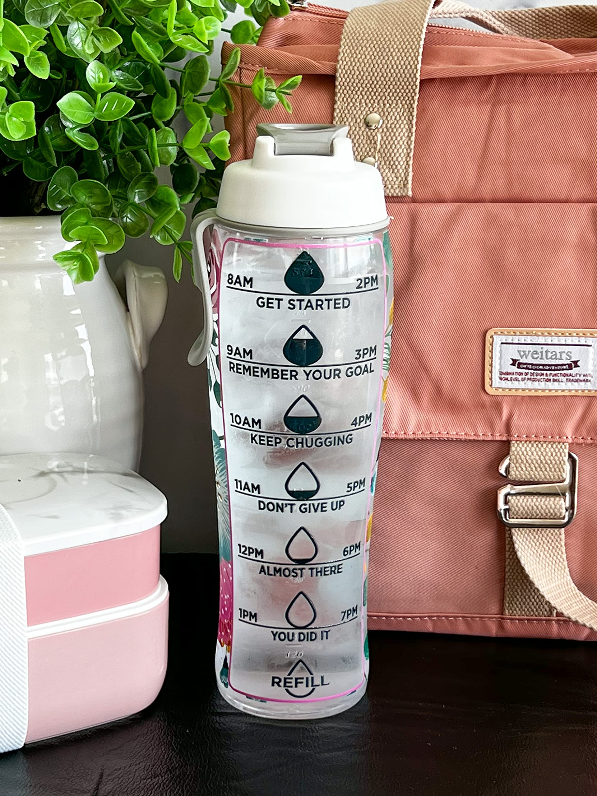Motivational water bottle with text and measurements on a surface with a pink bag and plant in the background.